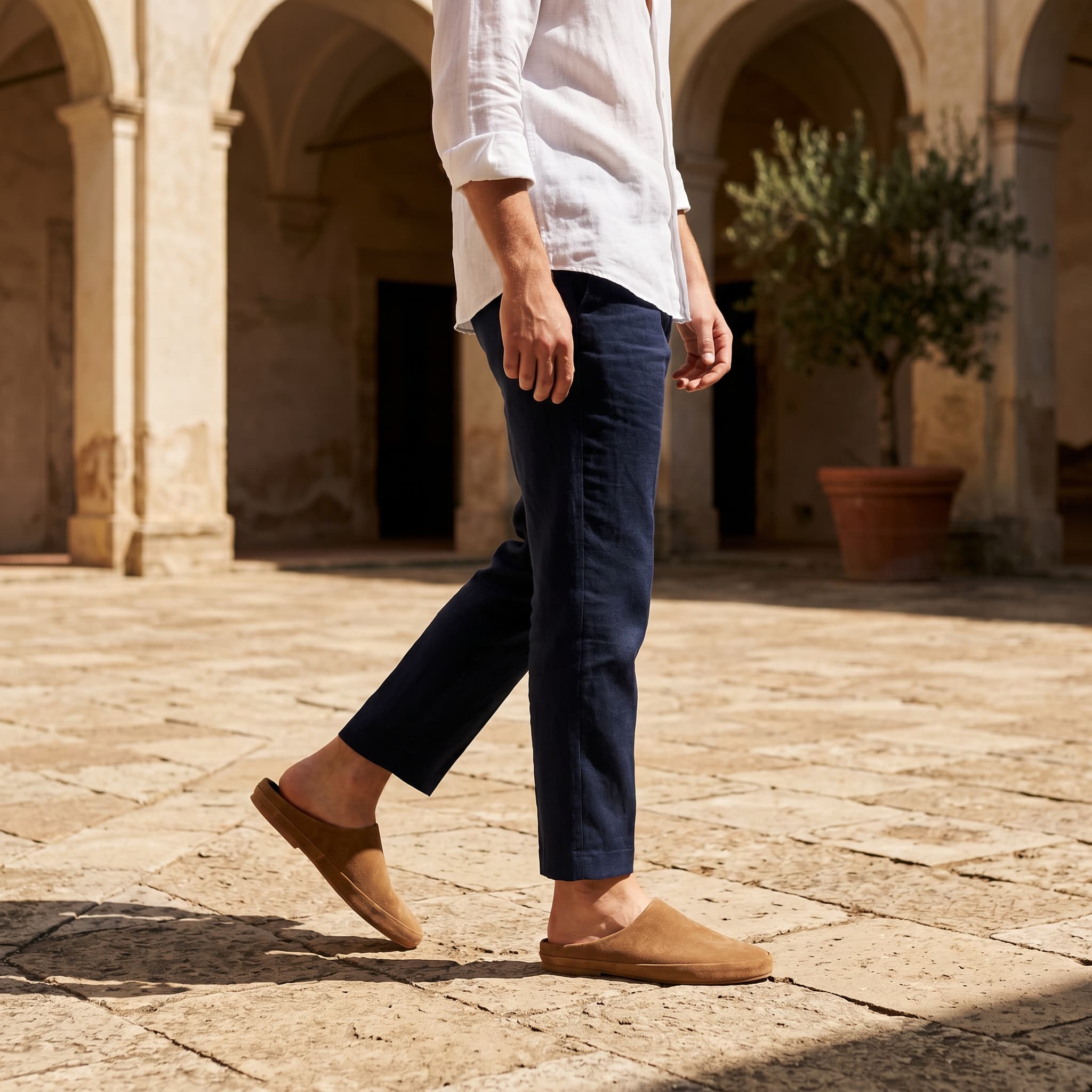 Person wearing navy pants and brown shoes walking on a stone pavement with arches and plants in the background.