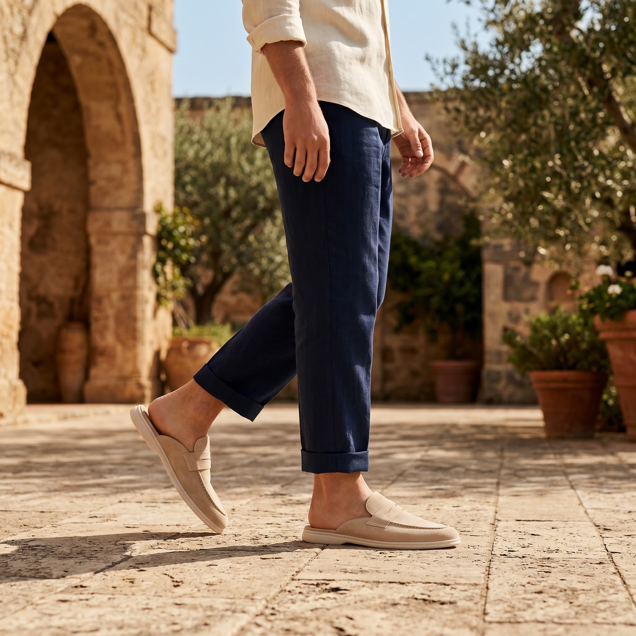 Person wearing navy pants and beige shoes walking on a stone path with a stone building and potted plants in the background.
