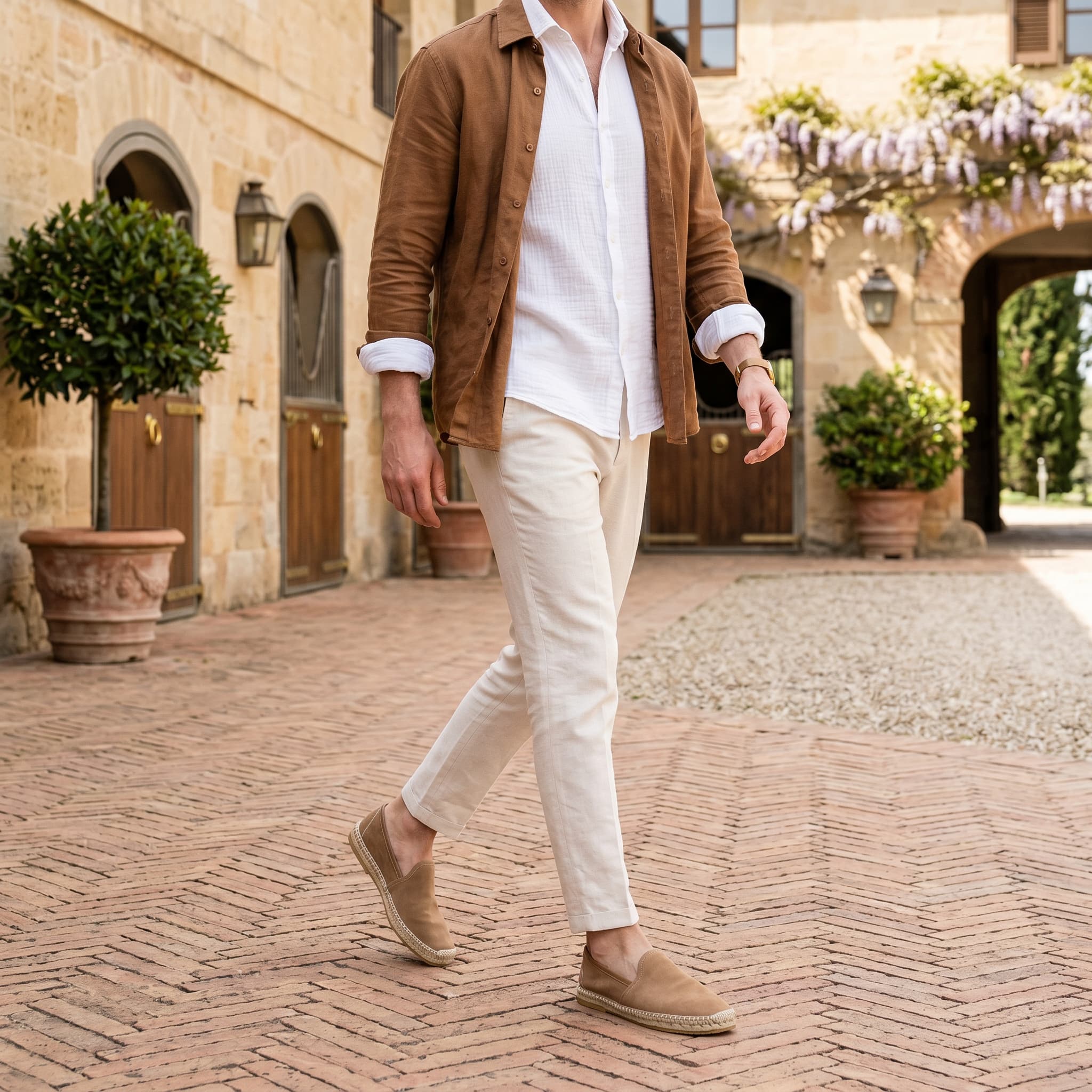 Man walking outdoors in a courtyard wearing a brown jacket, white shirt, beige pants, and tan shoes.