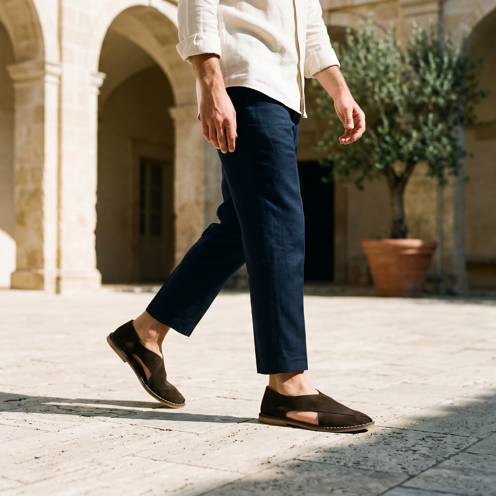 Person wearing navy pants and brown shoes walking on a stone pavement with a building and plant in the background.