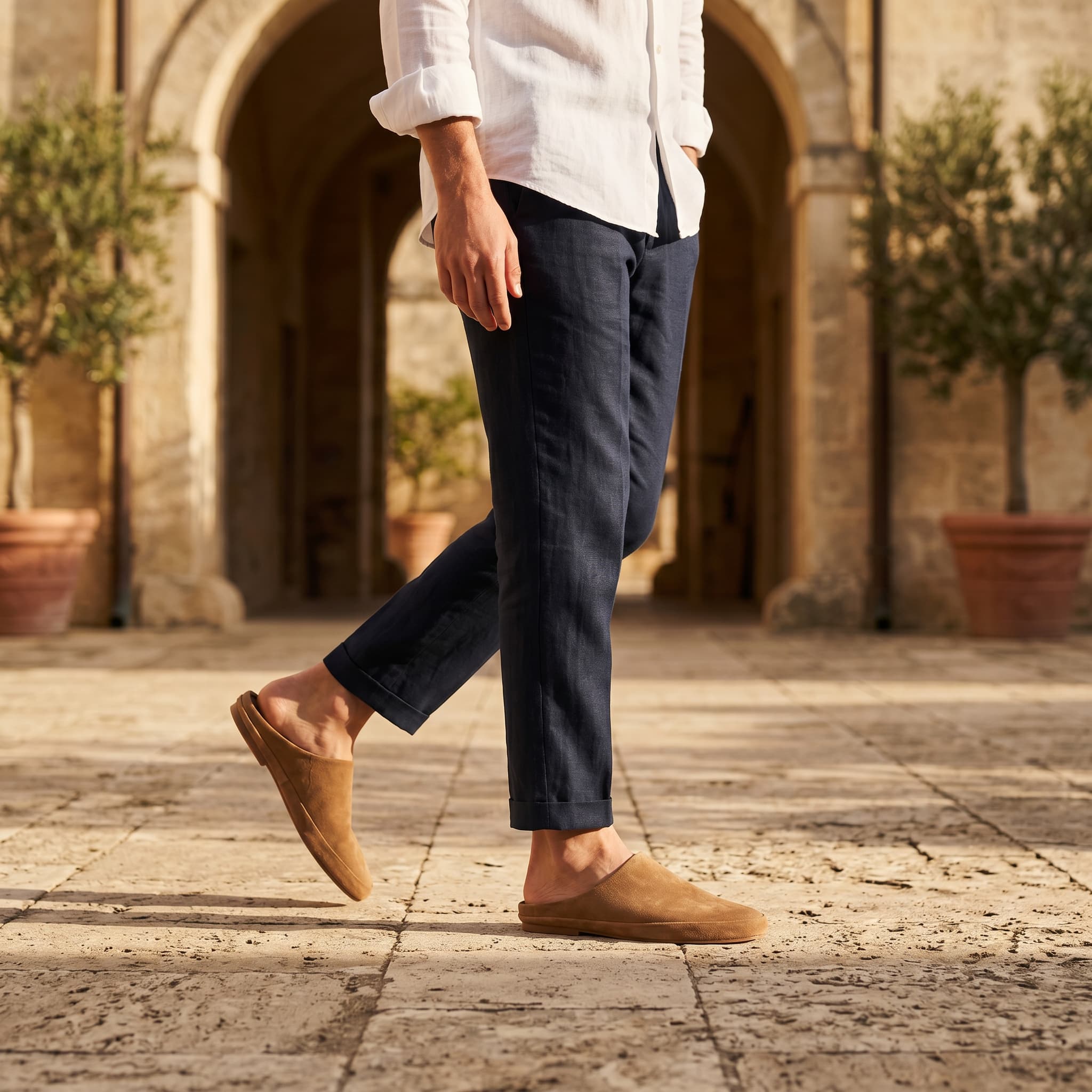 Person wearing brown shoes walking on a stone pavement with a stone building and potted plants in the background.